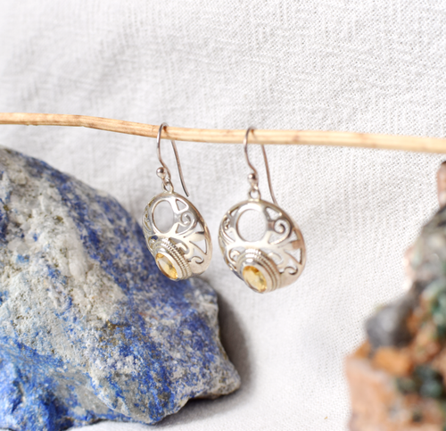 Silver earrings with a central gemstone hanging on a stick against a textured white background.
