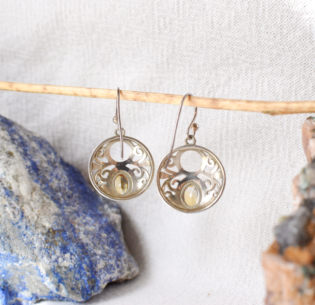 Silver earrings with intricate designs hanging on a stick against a white background.