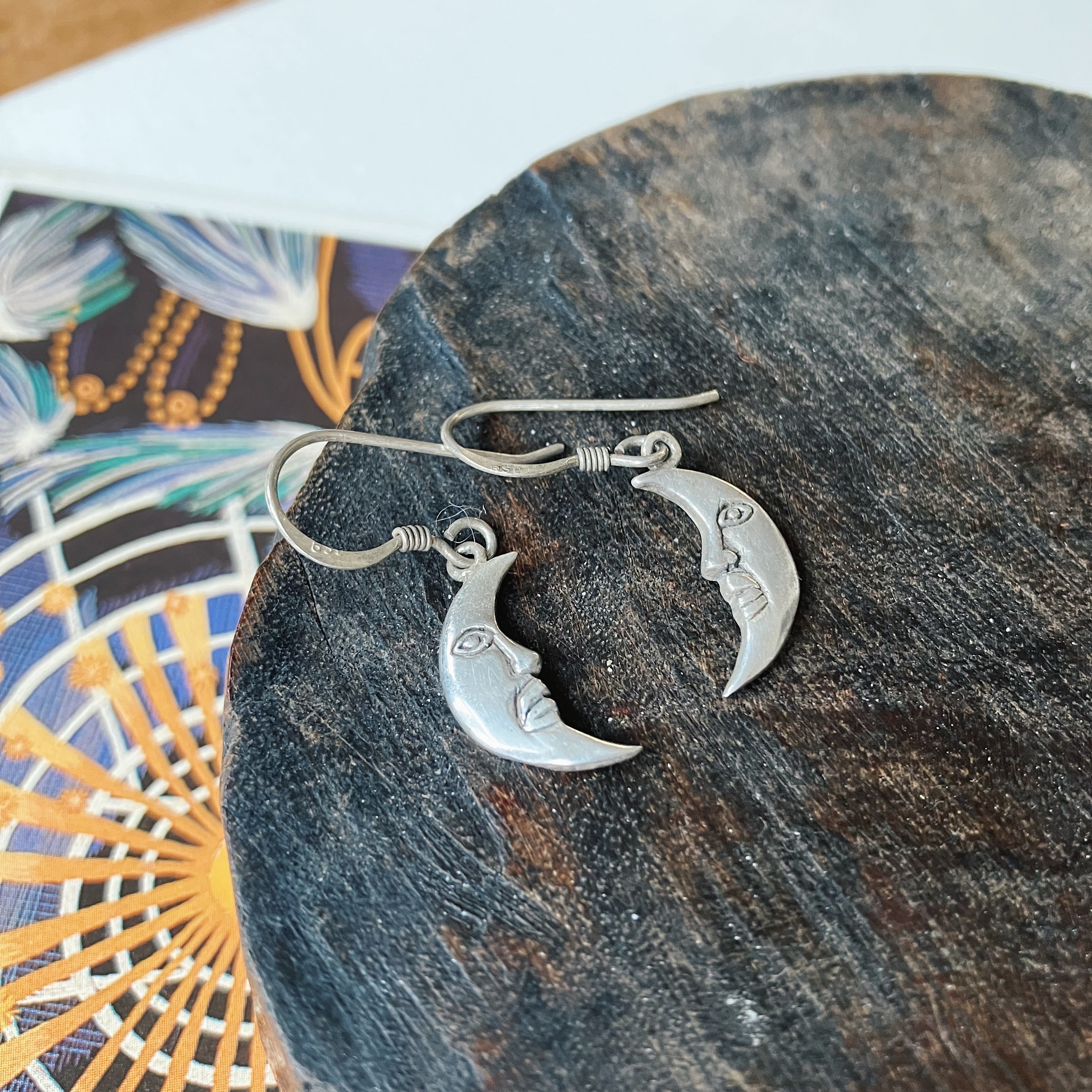 Silver crescent moon earrings on a dark stone surface with a colorful fabric background