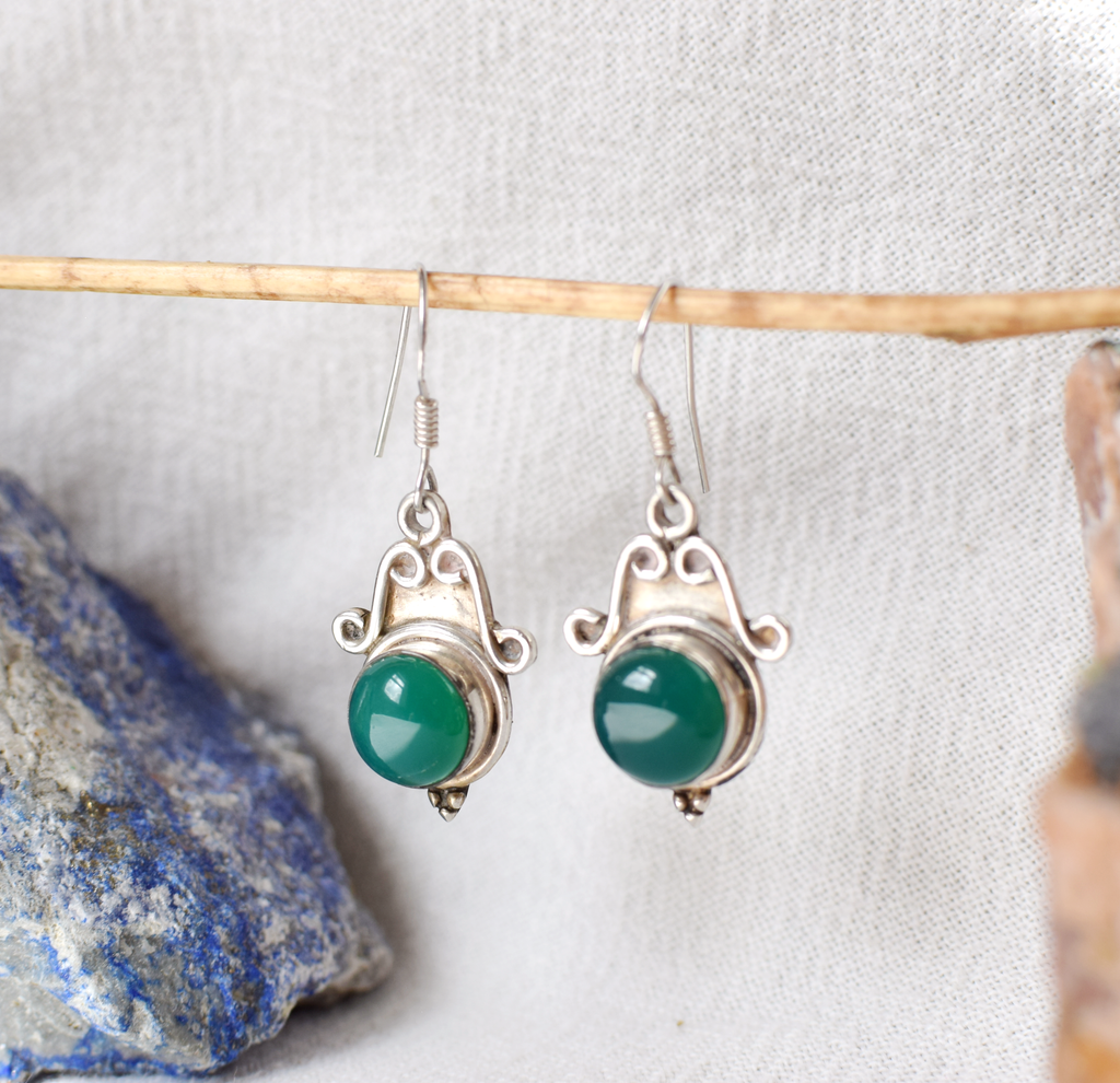 Silver earrings with green gemstones hanging on a wooden stick against a textured white background.