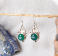 Silver earrings with green gemstones hanging on a wooden stick against a textured white background.